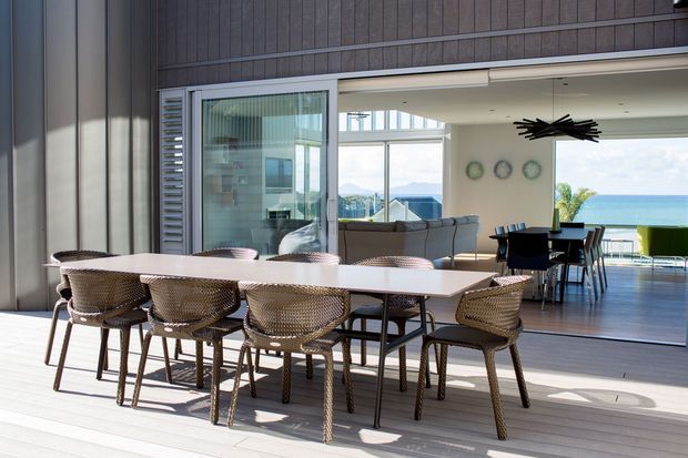A bright contemporary coastal dining area on a covered outdoor terrace, with a long dining table and woven chairs in the foreground. Large sliding glazed doors open to a connected living and dining space, with timber-look wall cladding, pale decking, modern lighting, and an ocean view beyond.