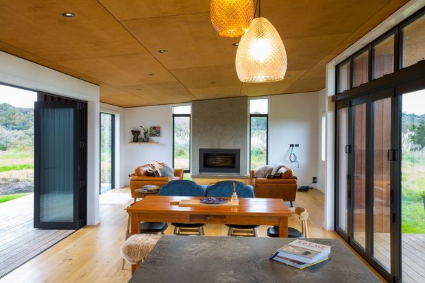 Open-plan living and dining area with a warm timber-panelled ceiling, light timber flooring, white walls, and extensive black-framed glazing. A central fireplace is set into a textured feature wall, with a timber dining table, upholstered seating, leather lounge chairs, and woven pendant lights. The room opens to an outdoor deck and landscaped surroundings.