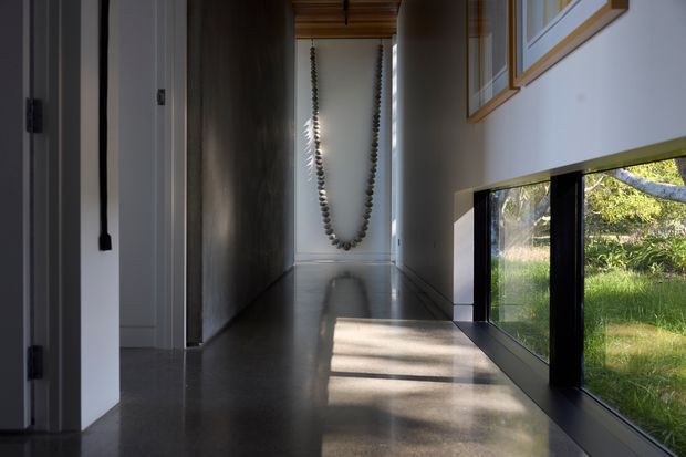 A narrow, minimalist interior hallway with a polished dark concrete floor, white walls, black-framed low horizontal glazing, and a timber-lined ceiling section. Sunlight enters through the glazing, creating strong shadows across the floor, while framed artwork and a hanging sculptural element add restrained visual interest. Garden vegetation is visible outside the windows.