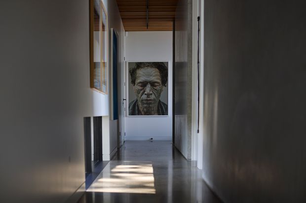 A long, narrow interior hallway with white walls, a dark polished floor, and a warm timber slatted ceiling. Framed artwork lines the walls, with a large portrait at the far end. Daylight enters from openings on the left, creating strong rectangular reflections across the floor. The corridor has a restrained, gallery-like contemporary character.