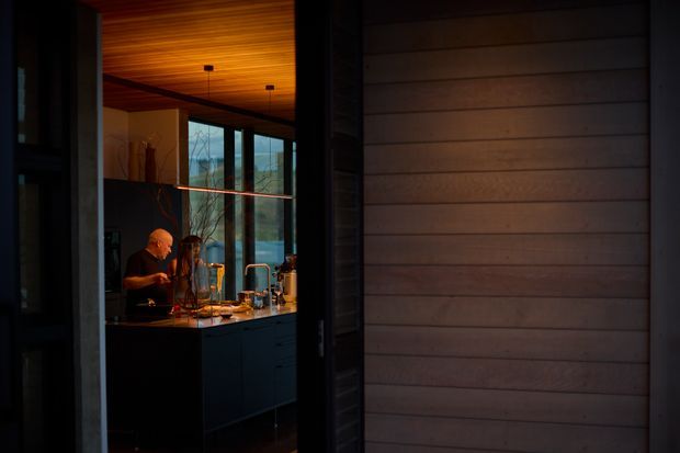 A moody view into a compact contemporary kitchen through a dark-framed opening. The kitchen features a dark island with integrated cabinetry, a warm timber-lined ceiling, a linear pendant or shelf light above the island, and large glazed windows overlooking a natural landscape. Two people are gathered at the island, with timber-framed horizontal cladding or panelling visible in the foreground.