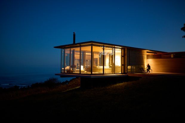 A modern single-storey coastal house photographed at dusk, with a long, low flat roof and extensive floor-to-ceiling glazing. Warm interior lighting reveals an open living area, while horizontal timber cladding extends toward a darker enclosed wing. The house sits on a sloping coastal site overlooking the water, with a projecting glazed corner and a sheltered outdoor seating area.