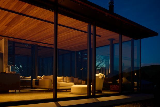 A contemporary glass-walled living room viewed from outside at dusk. The pavilion-like structure has extensive floor-to-ceiling glazing, dark slender framing, a broad flat roof, and a warm timber-lined ceiling. Inside are light upholstered sofas and ottomans arranged around a low coffee table, with warm interior lighting contrasting against the dark surrounding landscape.