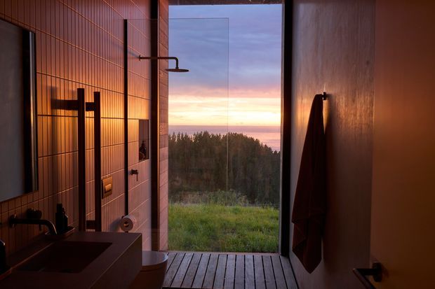 A moody modern bathroom with warm timber-toned tiled walls, a dark rectangular vanity and basin, black tapware, wall-mounted shower, and a timber-look floor. Full-height glazing opens to a tranquil forested landscape and sunset view, while a dark towel hangs on the right wall. The space has a refined, minimalist design with strong indoor-outdoor connection and dramatic warm lighting.