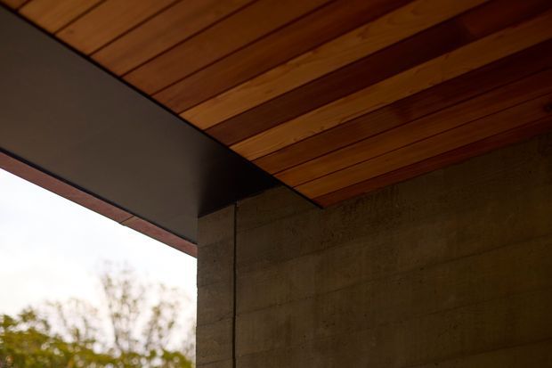 Close-up architectural detail of a covered exterior corner, showing a warm timber-lined soffit with exposed structural beams, a dark metal fascia, and a rough concrete block wall. Trees and bright sky are visible beyond the open edge.