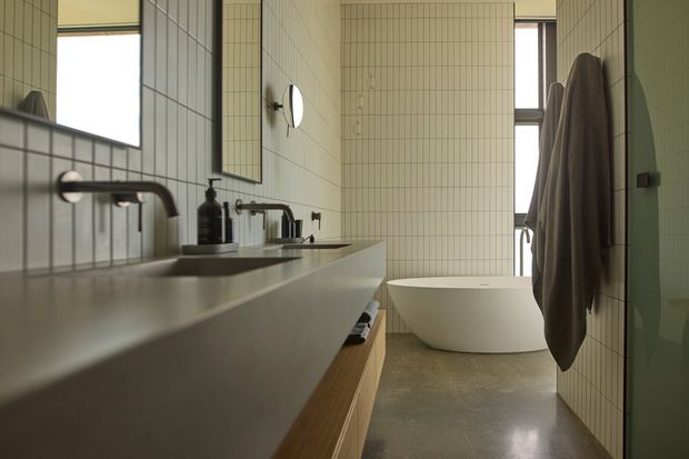 A narrow, contemporary bathroom with a long floating vanity and integrated pale benchtop, twin wall-mounted faucets, rectangular white tiled walls, and timber open shelving beneath the vanity. A freestanding white bathtub sits at the far end beside tall windows. Dark towels hang on the right wall, while black fixtures and framed mirrors provide contrast. The room has a calm, softly lit, warm-neutral atmosphere.