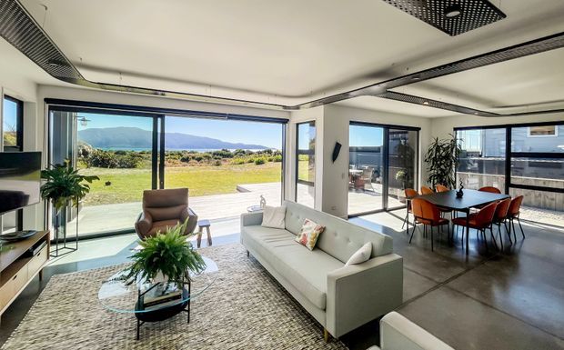 Bright contemporary open-plan living and dining area with full-height glazing overlooking a lawn and distant waterfront landscape. The room features polished concrete flooring, a pale sectional sofa, glass coffee table, large textured rug, indoor plants, and a dark dining table with orange dining chairs. Linear ceiling grilles and recessed lighting contribute to a clean modern fit-out.