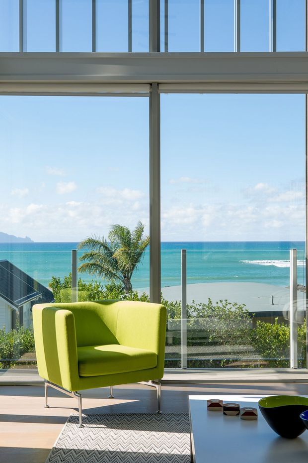 Bright contemporary living space with a lime-green upholstered armchair, low coffee table, patterned rug, and extensive floor-to-ceiling glazing. The glazing opens to a balcony or terrace with glass balustrades and expansive turquoise ocean and beach views. A palm tree and coastal landscape are visible outside.
