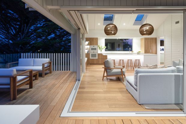 A bright open-plan living and kitchen area connected to a timber outdoor deck through fully retractable glazed doors. The interior features pale timber flooring, white walls and cabinetry, a kitchen island with timber stools, armchairs, white sofas, and two sculptural timber pendant lights. Skylights and broad openings create an airy indoor-outdoor connection, with a private deck and built-in seating visible outside.