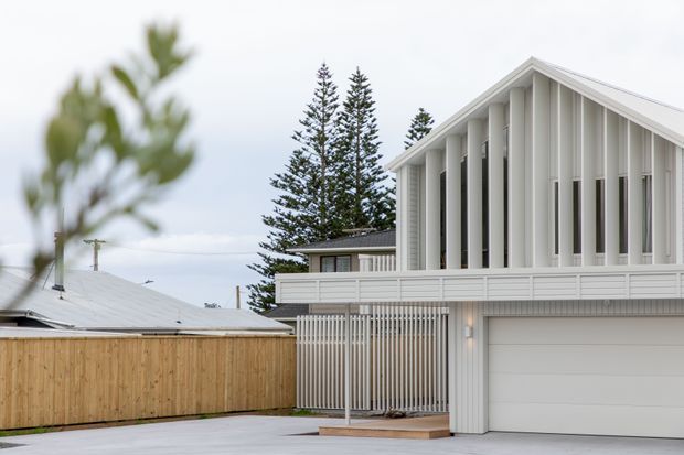 Exterior view of a contemporary two-storey home with a white vertical-screen facade, steep gabled roof, upper-level balcony, enclosed garage, and timber privacy fencing. The house sits in a suburban coastal-looking setting with tall evergreen trees in the background.
