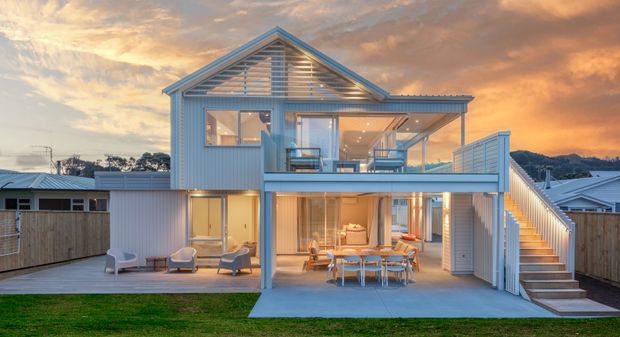 Exterior view of a contemporary multi-level coastal-style home at dusk, featuring a prominent gabled upper roof, horizontal louvres, pale vertical cladding, extensive glazing, elevated outdoor terraces, an open-plan dining and living area, and an external stair connecting the lower patio to the upper balcony.