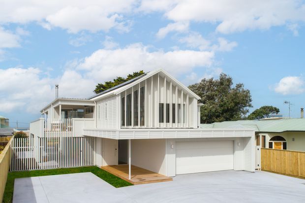 A bright contemporary multi-level white-clad house viewed from the street, featuring a prominent gabled upper volume with tall vertical windows, a broad lower-level garage, covered entry, timber decking, white privacy fencing, and a pale concrete driveway. The image shows a suburban coastal-style residential exterior under a partly cloudy sky.