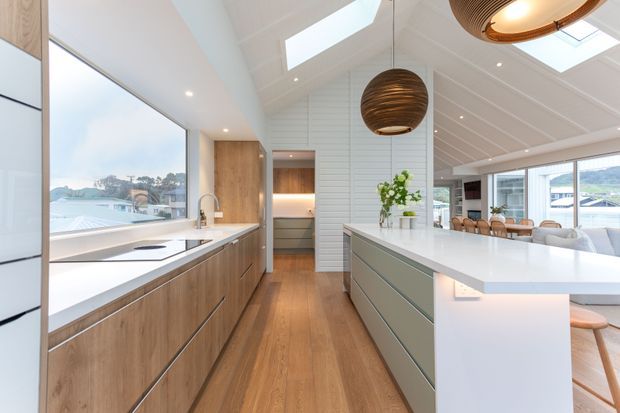 Bright contemporary kitchen with extensive timber cabinetry, a large white island, white shiplap-style wall panelling, timber flooring, skylights, and oversized woven pendant lights. The kitchen opens to a dining area with views through large windows toward a coastal or waterfront setting.