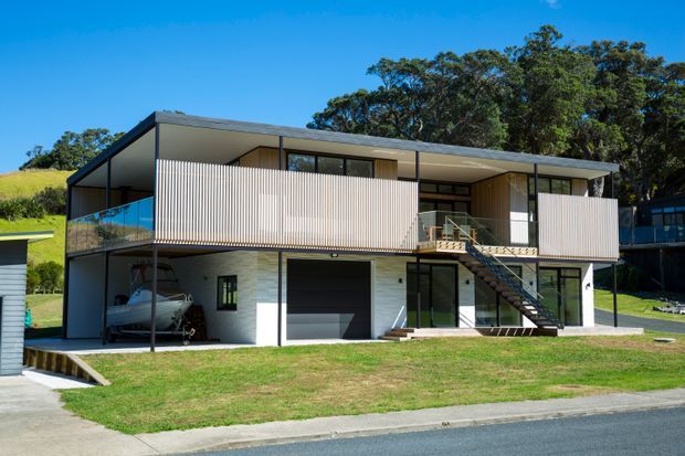 Exterior view of a contemporary two-storey residential house on a sloping suburban site. The building has a low-pitched roof, dark structural framing, light vertical privacy screens and cladding, extensive glazing, an upper-level balcony, and an external steel stair leading to the main entry. A garage and covered carport are visible at ground level.