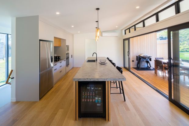 Bright open-plan kitchen with a long stone island, integrated sink and under-counter beverage fridge, stainless-steel appliances, light timber flooring, white cabinetry, and extensive black-framed sliding glass doors opening to a timber deck and garden. A dining area is visible beyond the glazing.