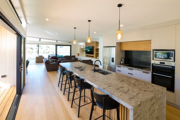 A bright, open-plan contemporary kitchen and living area. A large waterfall-edge stone island with seating for several people dominates the foreground, paired with white built-in cabinetry, a dark horizontal-tile splashback, timber-look overhead joinery, integrated appliances, and pendant lighting. Light timber flooring extends toward a relaxed living space and expansive glazed doors at the rear.