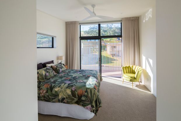 A bright contemporary bedroom with a low bed dressed in a bold tropical botanical bedspread, neutral carpet, full-height beige curtains, and a vivid lime-green upholstered accent chair. Large glazed doors open to a small balcony or terrace with vertical screening, while a ceiling fan and simple white walls create an airy, relaxed atmosphere.