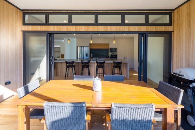 A bright outdoor dining terrace looks through wide dark-framed sliding doors into an open-plan kitchen. The terrace has a large timber dining table with woven outdoor chairs and a barbecue, while the kitchen features a timber island with bar seating, dark cabinetry, and stainless-steel appliances. Timber wall cladding and a row of high clerestory-style windows frame the opening.