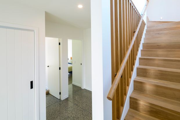 Bright contemporary interior view of a hallway and stairway. The image shows white walls and doors, grey carpeted flooring in the hall, and a timber staircase with matching vertical timber balustrade and handrail. An open doorway provides a partial view into a bedroom.