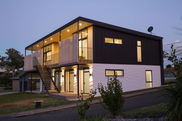 A contemporary two-storey residential building photographed at dusk, with a strongly rectilinear form, dark vertically ribbed upper cladding, white masonry lower walls, extensive glazing, projecting balconies, and an external steel stair. Warm interior lighting contrasts with the cool blue evening sky. The building sits in a suburban landscaped setting with a paved driveway and small garden areas.