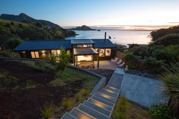 Elevated exterior view of a modern coastal home set into a lush, sloping site overlooking a sheltered bay. The low-profile dark metal roof, extensive glazing, timber-toned interiors, stepped concrete landscape, and illuminated stairway are clearly visible. Native-style planting surrounds an outdoor terrace, with mountainous coastline and water in the background at sunset.