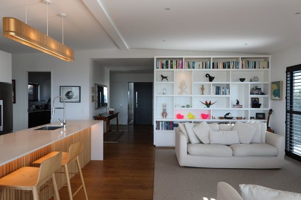 Open-plan contemporary living, dining and kitchen area with a warm timber floor, pale walls and a large white built-in shelving unit filled with books and decorative objects. A white sofa faces the shelving, while the kitchen island in the foreground has a light stone benchtop, white cabinetry and a timber slatted feature front, paired with upholstered bar stools. A linear timber pendant hangs above the island, and dark-framed glazing with horizontal blinds is visible on the right.