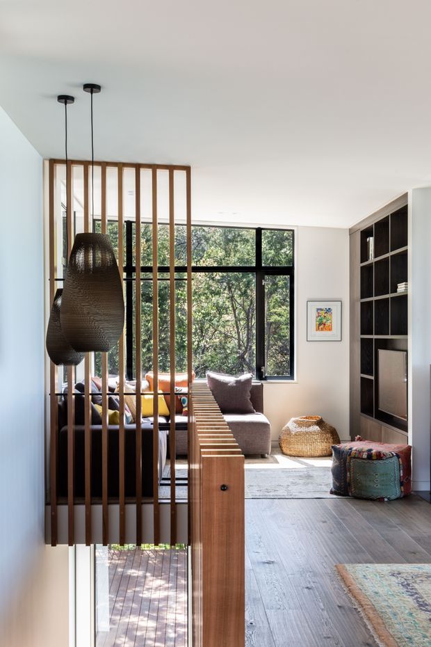 A bright contemporary living room viewed from an entry or circulation area, with full-height black-framed windows overlooking trees, a grey sectional sofa, colourful cushions, woven pendant lighting, timber vertical slat screening, and a dark built-in shelving and media unit. Warm timber flooring and natural woven accents create a relaxed, refined interior.