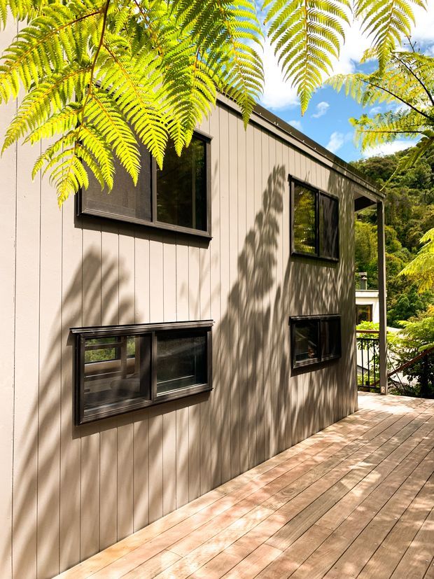 Exterior view of a compact multi-storey dwelling set in a leafy, sloped landscape. The facade is clad in vertically grooved light taupe weatherboard-style panels with dark-framed rectangular windows. A timber deck runs alongside the building, while lush fern foliage partially frames the scene. The roof appears low-pitched and the overall design is simple and contemporary.