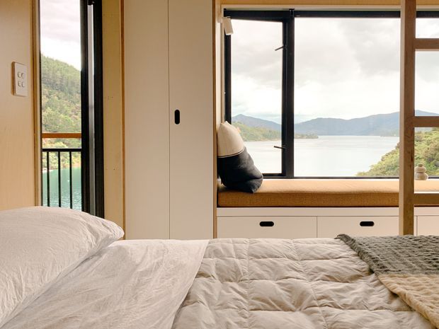 A calm, compact bedroom with a bed in the foreground and a built-in window seat overlooking a turquoise inlet and wooded mountainous landscape. The room features pale timber-toned wall and cabinetry, a black-framed opening window, integrated drawers beneath the seat, and a small balcony visible through the glazing.