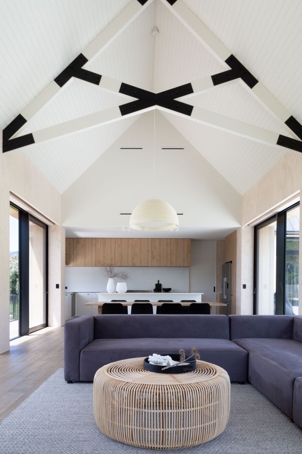 Open-plan living and dining area with a kitchen at the rear, featuring a dramatic vaulted white timber-lined ceiling with exposed black structural beams, pale timber wall finishes, large glazed openings, a dark sectional sofa, and a round woven timber coffee table.