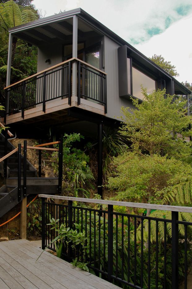 Exterior view of a compact modern multi-level dwelling set into lush vegetation on a sloped site. The building features dark grey vertical cladding, a low-pitched roof with broad eaves, large windows, timber decking, and black metal balustrades connected by exterior stairs and elevated walkways.