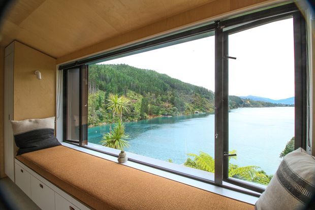 A compact, warm timber-lined window seat overlooking a turquoise lake and forested hills. The built-in bench includes drawers below, upholstered tan cushioning, neutral cushions, and large dark-framed operable glazing providing expansive water views.