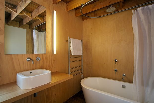 Compact rustic bathroom clad throughout in warm natural timber plywood, featuring a freestanding white bathtub with shower over bath, a white vessel basin on a timber vanity, exposed timber ceiling beams, a wall-mounted heated towel rail, a large mirror, and a slim vertical light. The space has an earthy, cabin-like character with visible exposed structure and a warm, low-key atmosphere.