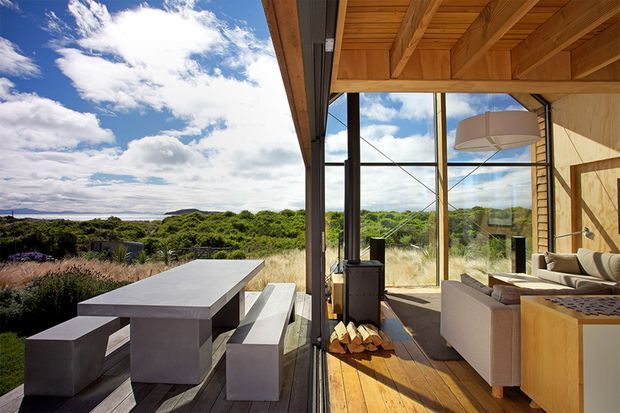 A contemporary indoor-outdoor living and dining area opening onto a deck, with full-height glazing framing a lush coastal landscape. The interior has a warm timber-lined ceiling and floor, exposed structural framing, a neutral lounge suite, and a built-in timber cabinet. Outside, a long concrete dining table with matching bench seating occupies the deck.