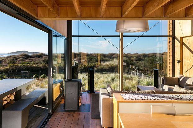 A bright contemporary living and dining space opening onto a coastal landscape through extensive floor-to-ceiling glazing. The room features warm timber ceiling beams and wall panelling, timber flooring, a built-in dining bench and table, upholstered seating, and a sculptural pendant light. Native dune vegetation and the ocean are visible beyond the glazing.