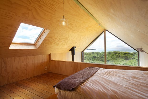 A warm timber-lined bedroom with a low sloping ceiling, exposed plywood wall and ceiling surfaces, timber floorboards, a skylight, and expansive triangular glazing overlooking a green coastal landscape. A bed occupies the foreground, with a small hanging bulb and simple wall-mounted lighting contributing to the rustic, cabin-like atmosphere.