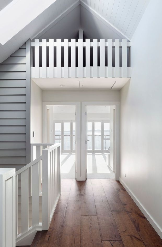 Bright interior view of an upper-level landing or hallway with a white-painted balustrade overlooking a stairwell. The space has a steep pitched, gabled ceiling with white vertical wall panelling, dark grey horizontal stair wall cladding, white interior doors, and warm medium-brown timber flooring. Sunlight enters through glazed doors at the end, creating a clean, airy atmosphere.