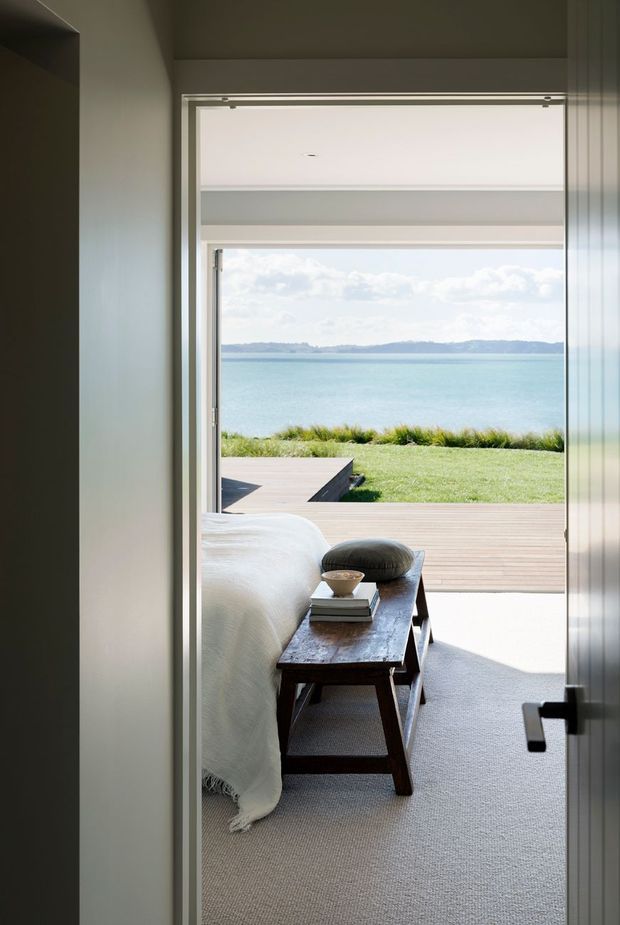A calm bedroom viewed through an open doorway, with a neatly made bed, dark timber bench at the foot, neutral carpeting, and minimalist white walls. Full-height glazing opens the room toward a timber deck, lawn, and a bright turquoise waterfront landscape beyond.