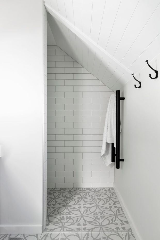 A compact bathroom beneath a sloped ceiling, featuring white vertical wall panelling, a white subway-tile feature wall, patterned grey-and-white floor tiles, matte black hooks, and a black heated towel rail with a white towel. The space is bright, clean, and highly tailored to the under-stair or attic-like geometry.
