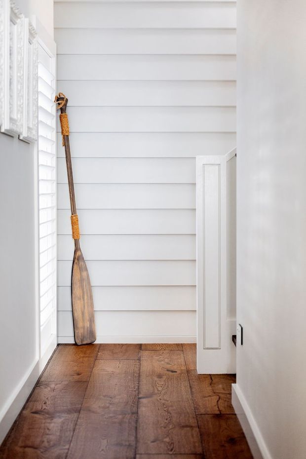 Bright narrow interior passage with white horizontal wall panelling, plantation shutters, ornate white trim, and warm medium-brown timber flooring. A decorative wooden paddle leans against the wall, and a partially open white door leads further into the home.