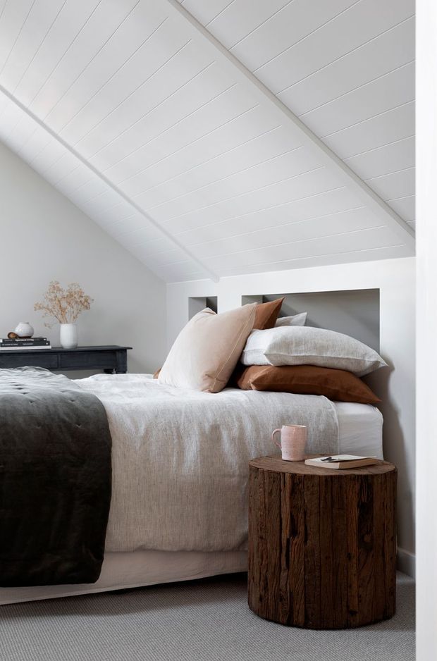 A calm, contemporary bedroom with a low-profile bed dressed in layered white, beige, grey, and rust-brown textiles. The room has a white pitched, panelled ceiling, pale walls, a recessed bedhead niche, grey carpet, a dark timber console, and a rustic cylindrical tree-trunk bedside table. Soft natural light creates a warm, understated atmosphere.
