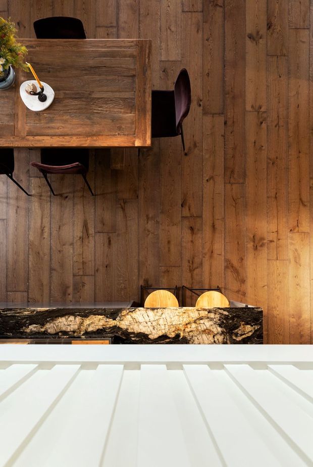 A top-down architectural interior photograph of a warm dining and kitchen area. A rustic solid timber dining table with dark dining chairs occupies the upper portion, while a dramatic dark stone kitchen island with prominent natural veining spans the lower middle. Vertical timber wall panelling and timber flooring create a strongly wood-lined setting, with bright linear ceiling or roof elements visible in the foreground.
