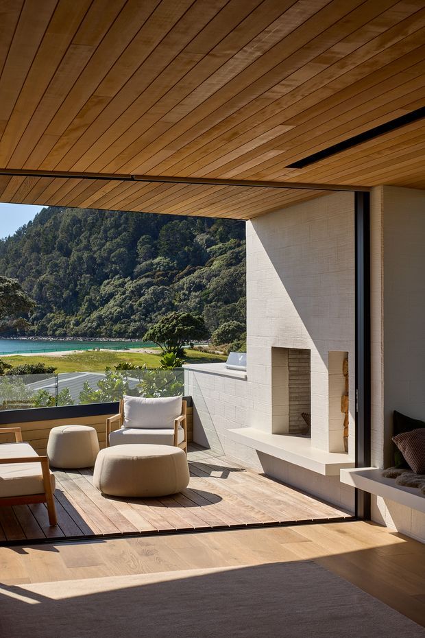 A bright, open-plan indoor-outdoor living space with a timber-lined ceiling, pale tiled fireplace wall, and large retractable glazed opening onto a timber deck. The room contains a built-in window seat, upholstered lounge chairs and ottomans, and a large neutral rug. Beyond the deck is a turquoise body of water and densely forested hills. The design has clean contemporary lines and a warm natural material palette.