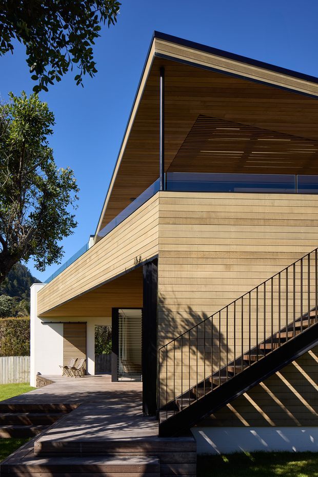 Exterior architectural photograph of a contemporary multi-level residence with extensive horizontal timber cladding and timber soffits. The composition highlights a sharply pitched gable roof, deep cantilevered upper volume, recessed outdoor terrace, dark metal-framed glazing and an external black metal staircase. Timber decking and landscaped surroundings are visible under clear daylight.