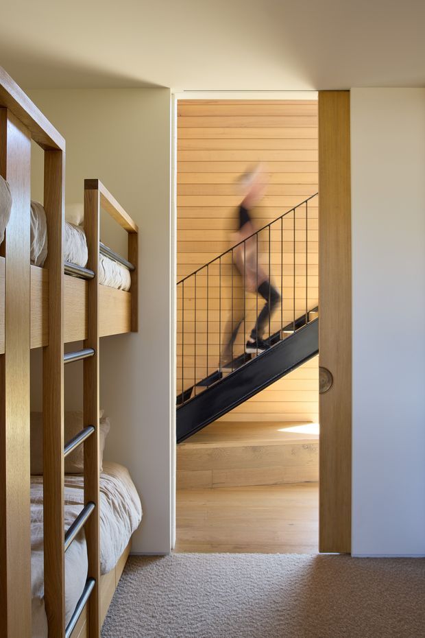 A compact bunk bedroom viewed toward an open doorway leading to a stairwell. Built-in timber bunk beds with white bedding line the left side, while the adjoining stair features warm horizontal timber wall cladding, a black metal balustrade, and timber flooring. The room has soft neutral walls, carpet flooring, and a simple contemporary fit-out. A blurred person is moving on the stairs.