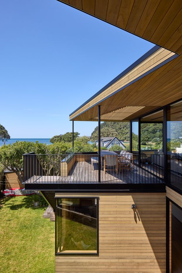 Exterior architectural photograph of a contemporary multi-level coastal home with extensive timber cladding and soffits, dark slender metal framing, and large glazed openings. The image focuses on an elevated outdoor terrace with black metal balustrades, dining furniture, and a slatted timber pergola/ceiling. Ocean, landscaped gardens, and neighbouring rooflines are visible beyond the deck.
