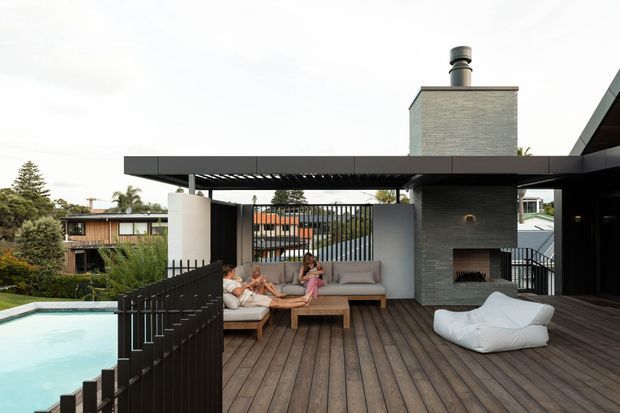 A contemporary rooftop-style outdoor entertaining deck with dark timber decking, a swimming pool, black metal balustrades, a covered pergola, built-in seating, and a substantial outdoor fireplace with chimney. The space overlooks neighbouring homes and landscaped surroundings, with a relaxed lounge arrangement and pale outdoor beanbag.