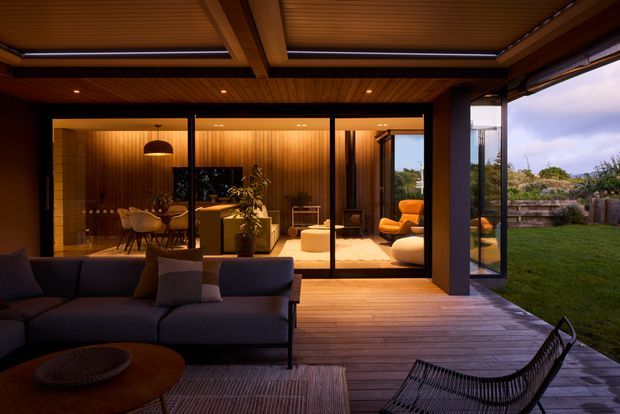 A sophisticated indoor-outdoor living area viewed from a covered timber deck at dusk. The foreground contains a dark upholstered sofa, round timber coffee table and woven lounge chair, while floor-to-ceiling glazing opens onto a warm, wood-lined interior living room with additional seating and a pendant light. A landscaped lawn is visible beyond the open glass doors. The design features strong indoor-outdoor connection, timber decking and ceiling lining, dark structural framing, and warm concealed lighting.