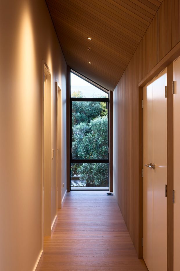 A narrow, warm-toned residential hallway with timber flooring, a sloped ceiling clad in narrow timber boards, and a vertical timber feature wall on the right. Plain light-painted walls and several flush interior doors line the corridor. A tall black-framed glazed window at the end provides abundant natural light and views of dense greenery outside, while recessed ceiling lights add warm illumination.