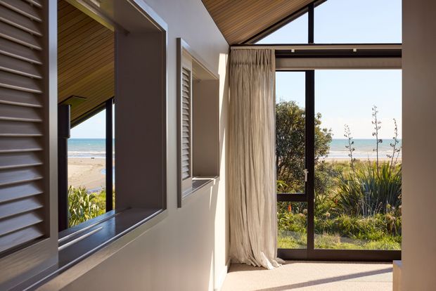 A bright coastal interior corner with floor-to-ceiling dark-framed glazing opening toward a sandy beach and ocean. The room has white plaster walls, louvred window shutters, a warm timber-lined sloping ceiling, and full-height cream curtains. Sunlight creates a calm, airy atmosphere, with coastal landscaping visible outside.